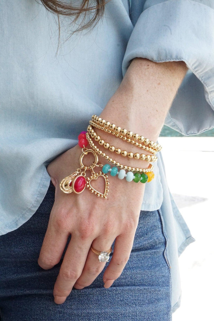 Hand wearing multiple gold bracelets with colorful charms against a light blue shirt.