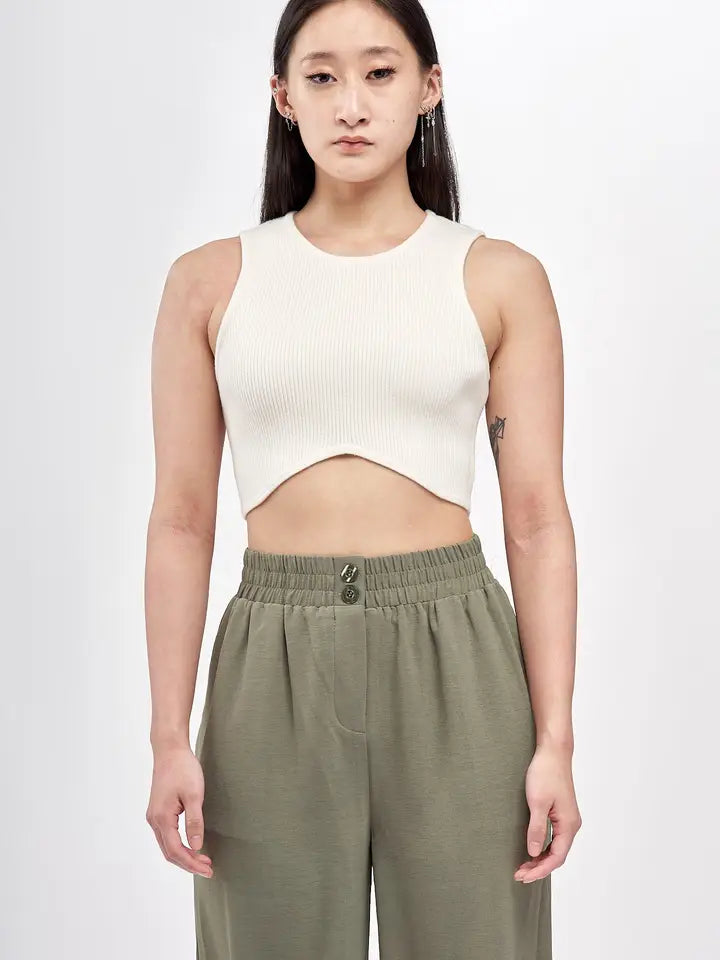 A woman stands facing forward, wearing a fitted, ribbed white crop top with a rounded hemline that dips at the center. She pairs it with high-waisted olive green pants featuring an elastic waistband and double-button closure.