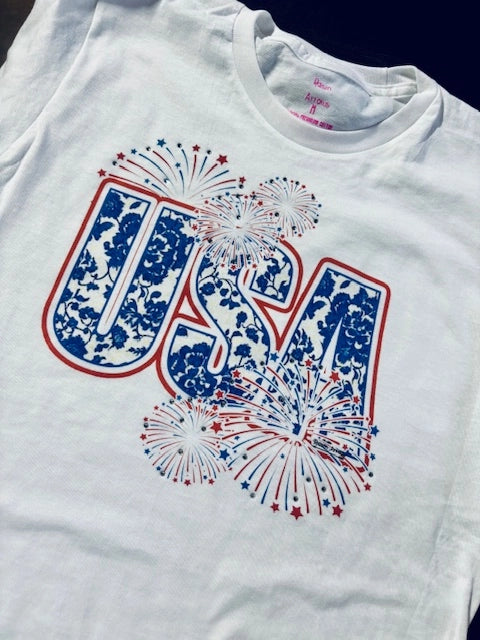White graphic t-shirt with red and blue “USA” text and sparkling firework design accented with jewels.

