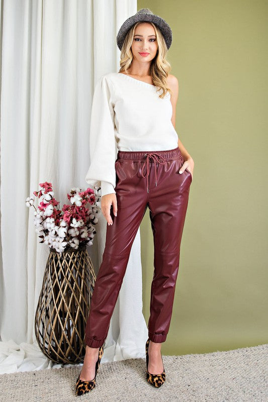 A close-up shot of a woman wearing a pair of wine-colored faux leather jogger pants with a drawstring waist and elastic cuffs. Her hands are in the side pockets, and she is wearing a white top.